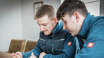 Zwei Männer in blauen Arbeitsjacken sitzen an einem Tisch und schauen konzentriert auf ein Dokument oder ein Gerät.
