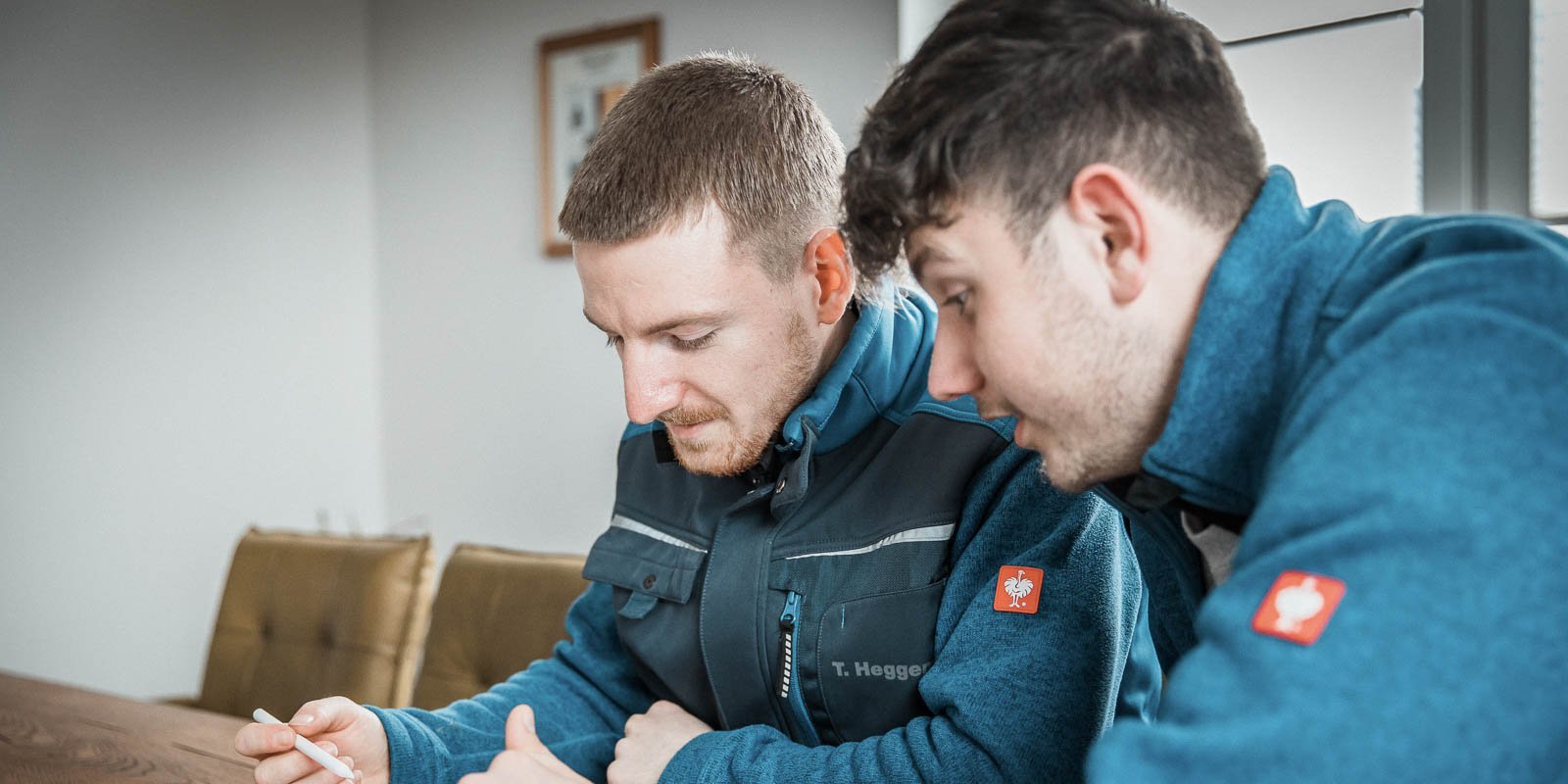 Zwei Männer in blauen Arbeitsjacken sitzen an einem Tisch und schauen konzentriert auf ein Dokument oder ein Gerät.