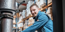 Ein lächelnder Mann in blauer Arbeitskleidung steht in einem Lager mit Regalen voller Kartons im Hintergrund.