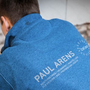 Ein Handwerker in einem blauen Pullover arbeitet konzentriert an einer Wand, während er Werkzeuge in der Hand hält.
