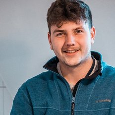 Der Mann trägt einen blauen Pullover und steht vor einer hellen Wand, während er mit einem breiten Lächeln in die Kamera schaut.