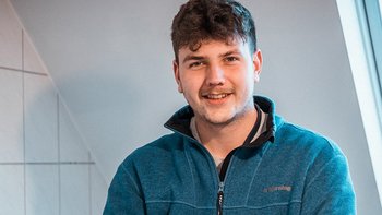 Der Mann trägt einen blauen Pullover und steht vor einer hellen Wand, während er mit einem breiten Lächeln in die Kamera schaut.