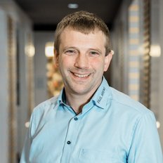 Der Mann mit einem freundlichen Lächeln trägt ein hellblaues Hemd und posiert in einem eleganten, gut beleuchteten Raum.