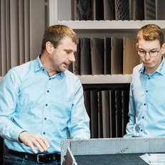 Zwei Männer in hellblauen Hemden betrachten Materialien in einem modernen Raum, während sie sich angeregt unterhalten.