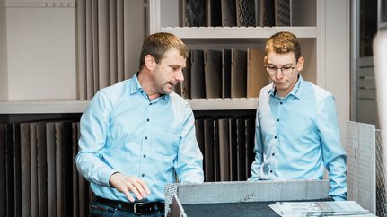 Zwei Männer in hellblauen Hemden betrachten Materialien in einem modernen Raum, während sie sich angeregt unterhalten.