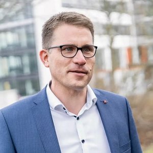 Ein Mann mit Brille und Anzug lächelt leicht, während er in einem modernen urbanen Umfeld steht.