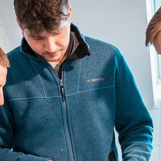 Drei junge Männer in blauen Jacken stehen zusammen und schauen konzentriert auf ein Gerät in ihren Händen.