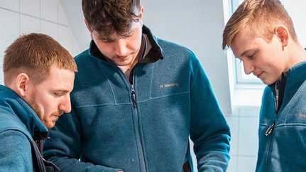 Drei junge Männer in blauen Jacken stehen zusammen und schauen konzentriert auf ein Gerät in ihren Händen.