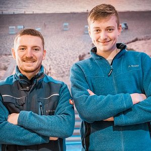 Zwei Männer stehen lächelnd nebeneinander, gekleidet in blaue Fleecejacken, vor einem Strandhintergrund.