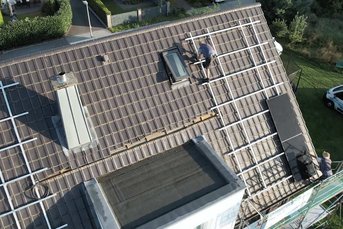 Ein Handwerker arbeitet auf einem Dach, während er eine Solaranlage installiert, umgeben von einem grünen Garten.
