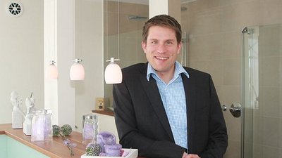 Ein lächelnder Mann in einem Anzug steht an einer modernen Theke mit dekorativen Objekten und sanfter Beleuchtung.