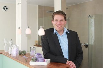 Ein lächelnder Mann in einem Anzug steht an einer modernen Theke mit dekorativen Objekten und sanfter Beleuchtung.