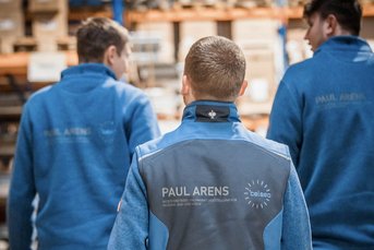 Drei Männer in blauen Arbeitsjacken gehen in einem Lager mit Regalen voller Kartons. Ihre Rückansicht ist sichtbar.