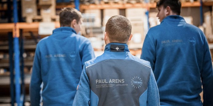 Drei Männer in blauen Arbeitsjacken gehen in einem Lager mit Regalen voller Kartons. Ihre Rückansicht ist sichtbar.