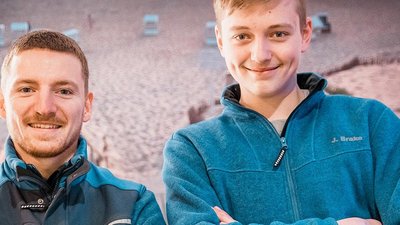 Zwei Männer lächeln freundlich in blauen Jacken, vor einem Hintergrund mit Strandstühlen und Sand.