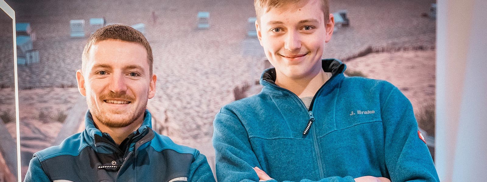 Zwei Männer lächeln freundlich in blauen Jacken, vor einem Hintergrund mit Strandstühlen und Sand.