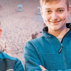 Zwei Männer lächeln freundlich in blauen Jacken, vor einem Hintergrund mit Strandstühlen und Sand.