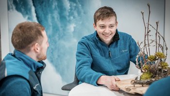 In einem modernen Raum unterhalten sich zwei Männer, einer lächelt und präsentiert ein kreatives Arrangement aus Holz und Pflanzen.