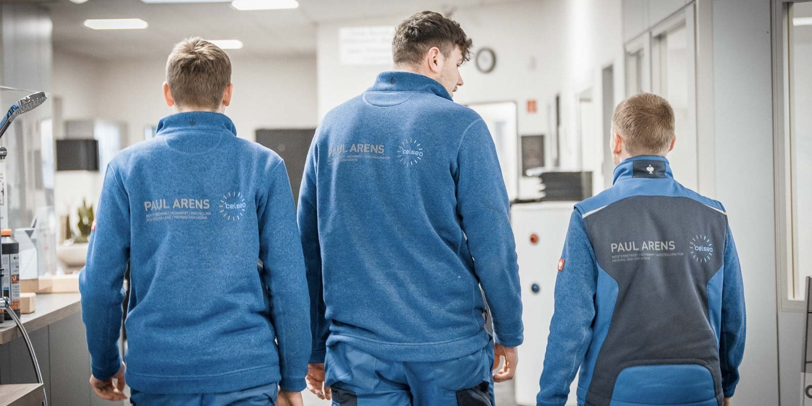 Drei Männer in blauen Arbeitsjacken mit dem Logo 'PAUL ARENS' gehen in einem modernen Bürogebäude.