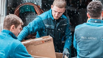 Drei Männer in blauen Arbeitsjacken laden einen Karton aus einem Lieferwagen, während es schneit.