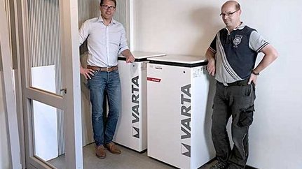 Zwei Männer stehen neben zwei weißen VARTA-Batteriespeichern in einem modernen Raum mit grauem Boden.