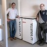 Zwei Männer stehen neben zwei weißen VARTA-Batteriespeichern in einem modernen Raum mit grauem Boden.