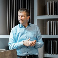 Ein Mann in einem hellblauen Hemd lächelt und gestikuliert, während er vor einer Wand mit verschiedenen Materialmustern steht.