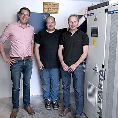 Ein VARTA-Gerät steht in einem Raum, während drei Männer lächelnd daneben stehen und eine positive Atmosphäre ausstrahlen.