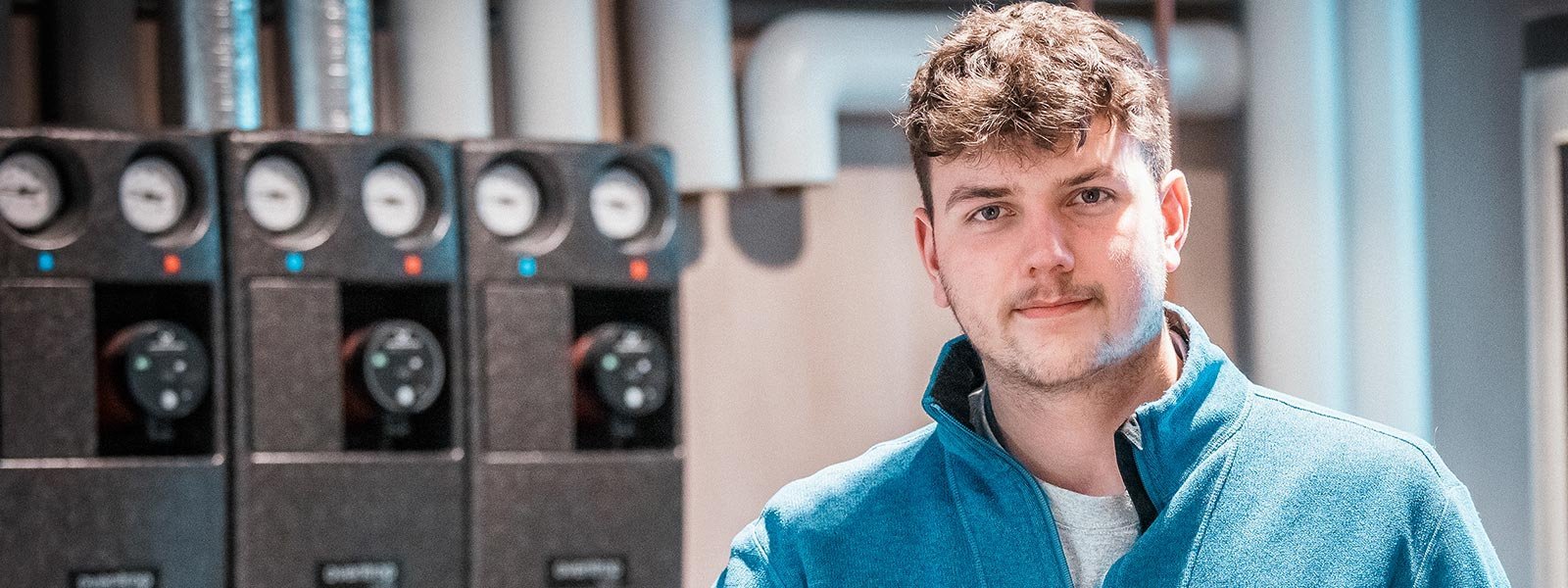 Ein junger Mann mit lockigem Haar trägt eine blaue Jacke und steht vor einer technischen Ausrüstung in einem modernen Raum.