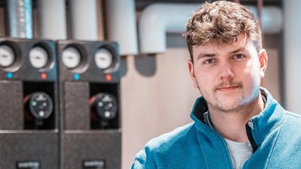 Ein junger Mann mit lockigem Haar trägt eine blaue Jacke und steht vor einer technischen Ausrüstung in einem modernen Raum.
