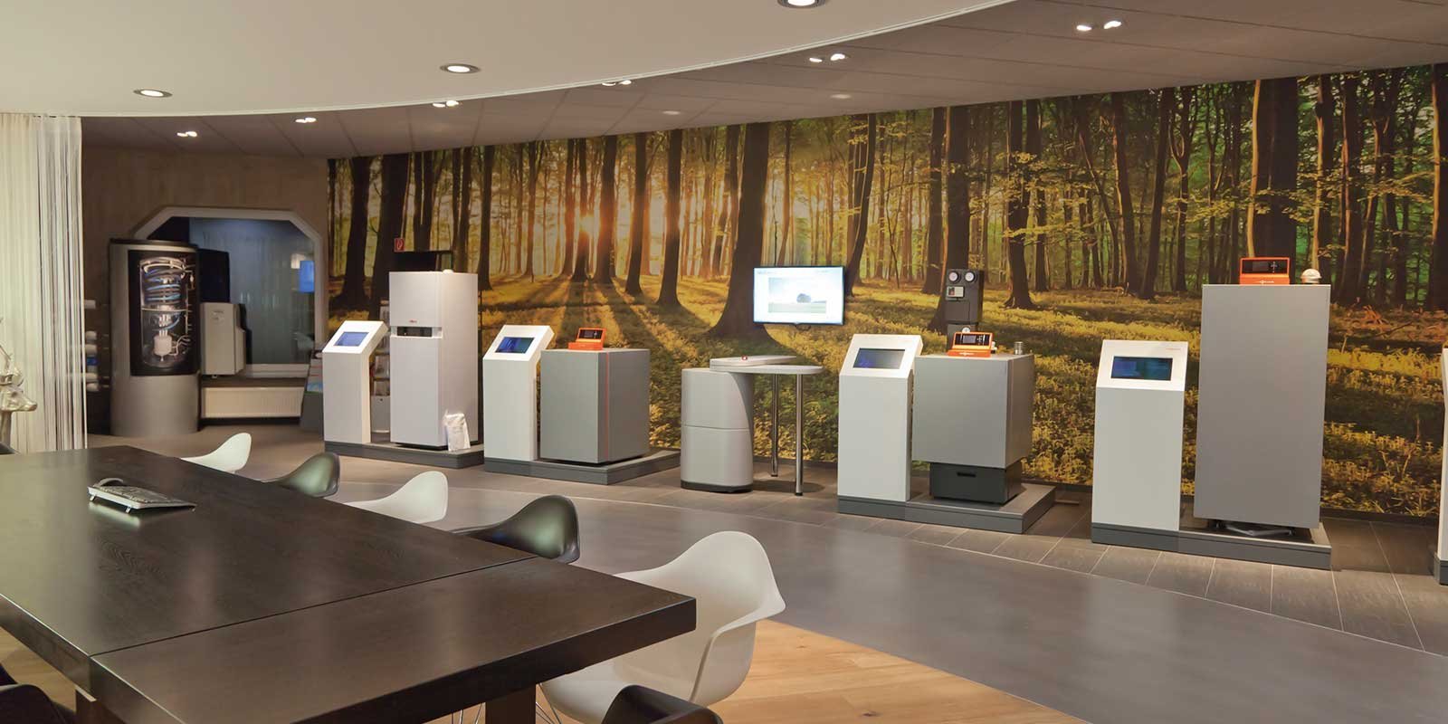 Ein moderner Ausstellungsraum mit mehreren interaktiven Geräten vor einer malerischen Waldtapete, die Sonnenstrahlen zeigt.