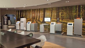 Ein moderner Ausstellungsraum mit mehreren interaktiven Geräten vor einer malerischen Waldtapete, die Sonnenstrahlen zeigt.