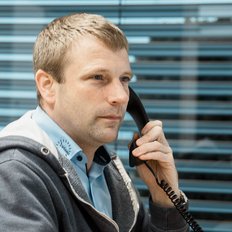 Ein Mann sitzt am Schreibtisch, telefoniert und schaut konzentriert aus dem Fenster mit Jalousien im Hintergrund.