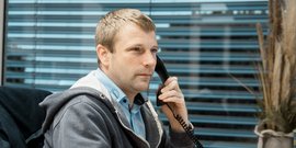 Ein Mann sitzt am Schreibtisch, telefoniert und schaut konzentriert aus dem Fenster mit Jalousien im Hintergrund.