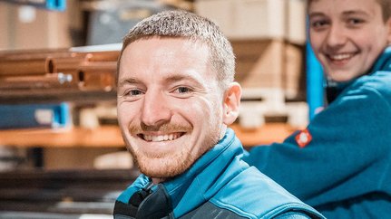 Zwei Männer in blauen Arbeitsjacken lächeln fröhlich in einem Lager mit Regalen und Rohren im Hintergrund.