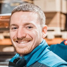 Zwei Männer in blauen Arbeitsjacken lächeln fröhlich in einem Lager mit Regalen und Rohren im Hintergrund.