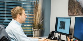 Ein Mann sitzt an einem Schreibtisch und schaut konzentriert auf zwei Computerbildschirme, während er an einem Projekt arbeitet.