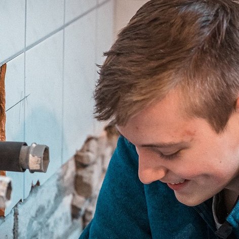 Ein junger Mann mit einem blauen Pullover arbeitet konzentriert an einer Wand mit freiliegenden Rohren und Fliesen.