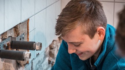 Ein junger Mann mit einem blauen Pullover arbeitet konzentriert an einer Wand mit freiliegenden Rohren und Fliesen.