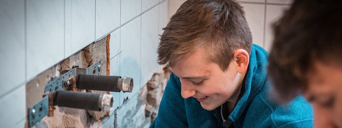 Ein junger Mann mit einem blauen Pullover arbeitet konzentriert an einer Wand mit freiliegenden Rohren und Fliesen.