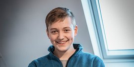 Ein lächelnder Jugendlicher trägt einen blauen Pullover und steht vor einem Fenster mit natürlichem Licht.
